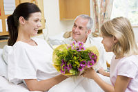 Picture of a female patient sitting up in a hospital bed and receiving flowers from an older man and young girl.